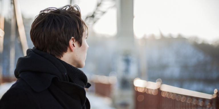 Man stands facing away from camera in a city area near a bridge.