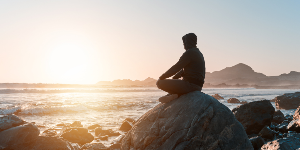 A man sits on a rock as the sun sets looking over the ocean.