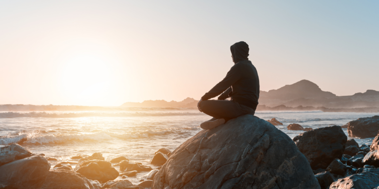 A man sits on a rock as the sun sets looking over the ocean.